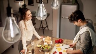 A woman watches a man cutting vegetable on a chopping board in a kitchen; you can see light bulbs in the upper part of the image
